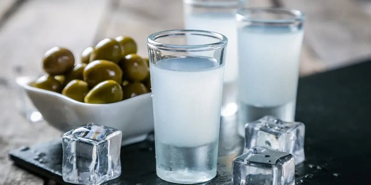 Trzy kieliszki ouzo z lodem i miseczka oliwek na drewnianym tle.