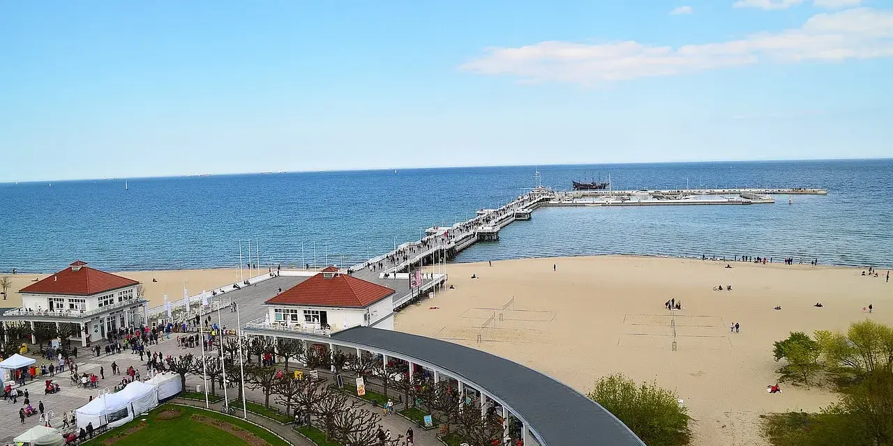 Sopot beach z molo, kawiarniami i spacerującymi ludźmi. W oddali widać statki.
