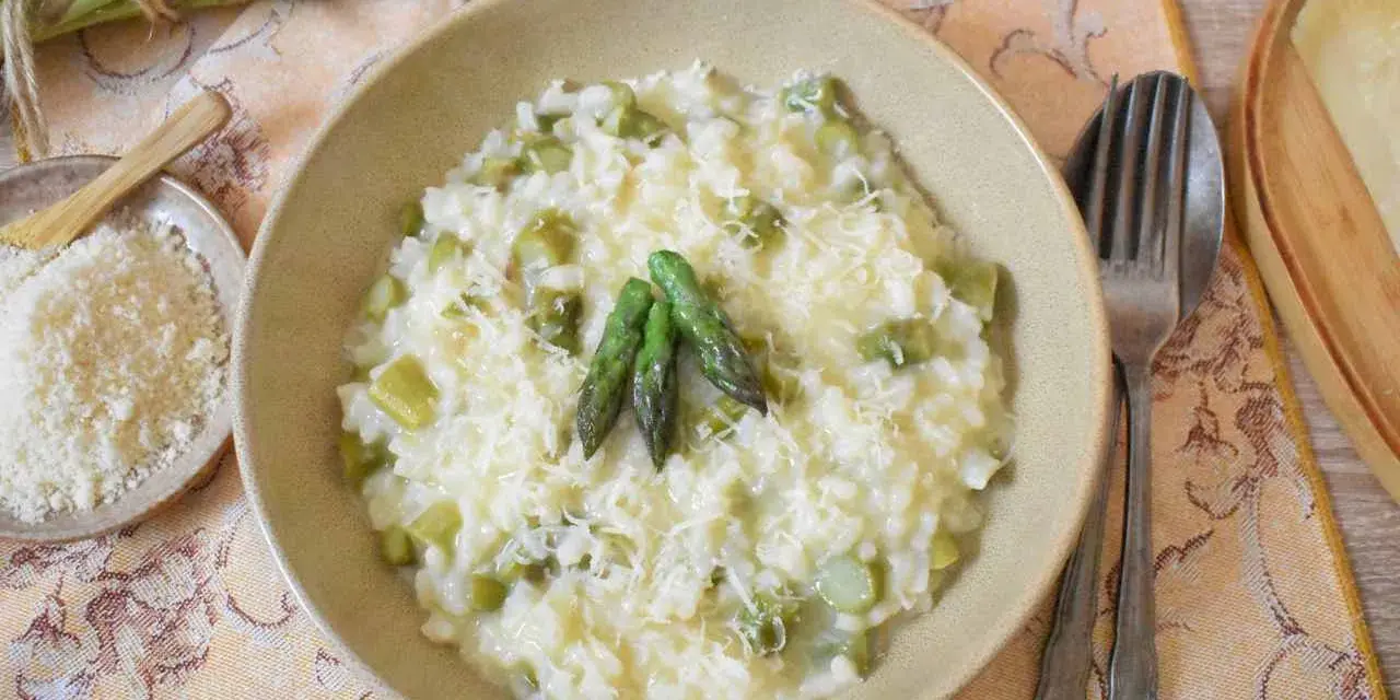 Kremowe risotto ze szparagami, posypane parmezanem, z trzema zielonymi szparagami na wierzchu. Obok miseczka z tartym serem i pęczek świeżych szparagów.