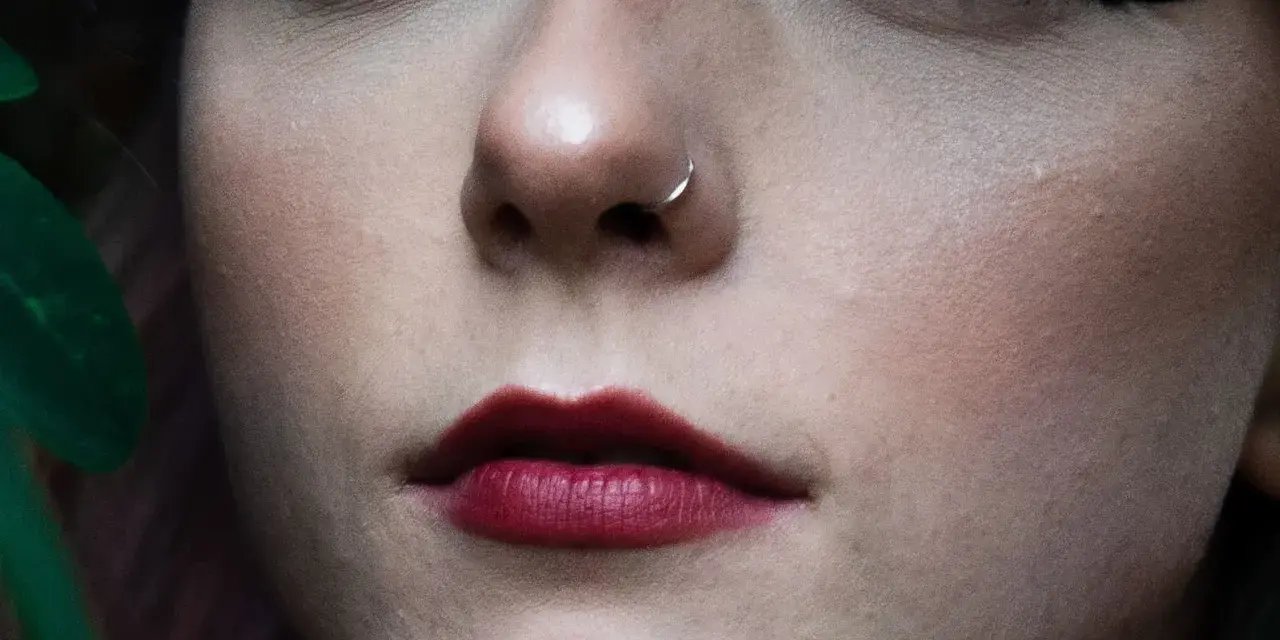 Delikatny kolczyk w nosie, jak rhino piercing, podkreśla pełne, ciemnoczerwone usta.