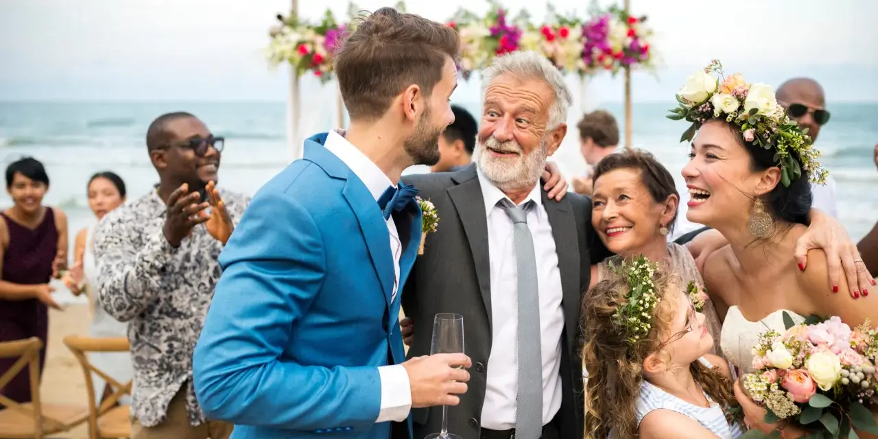 Szczęśliwy ojciec pana młodego w eleganckim garniturze śmieje się z synem na plażowym weselu.