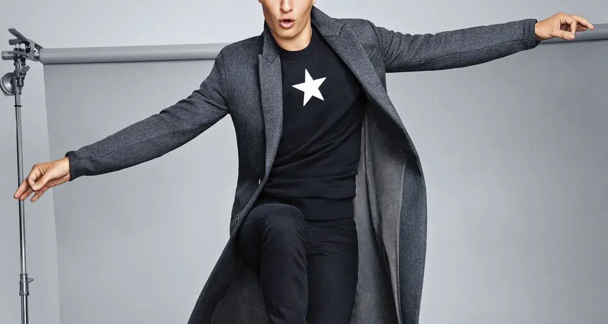 Joven con abrigo largo gris, camiseta negra con estrella blanca y vaqueros oscuros. Explora diversos tipos de abrigos para tu estilo.