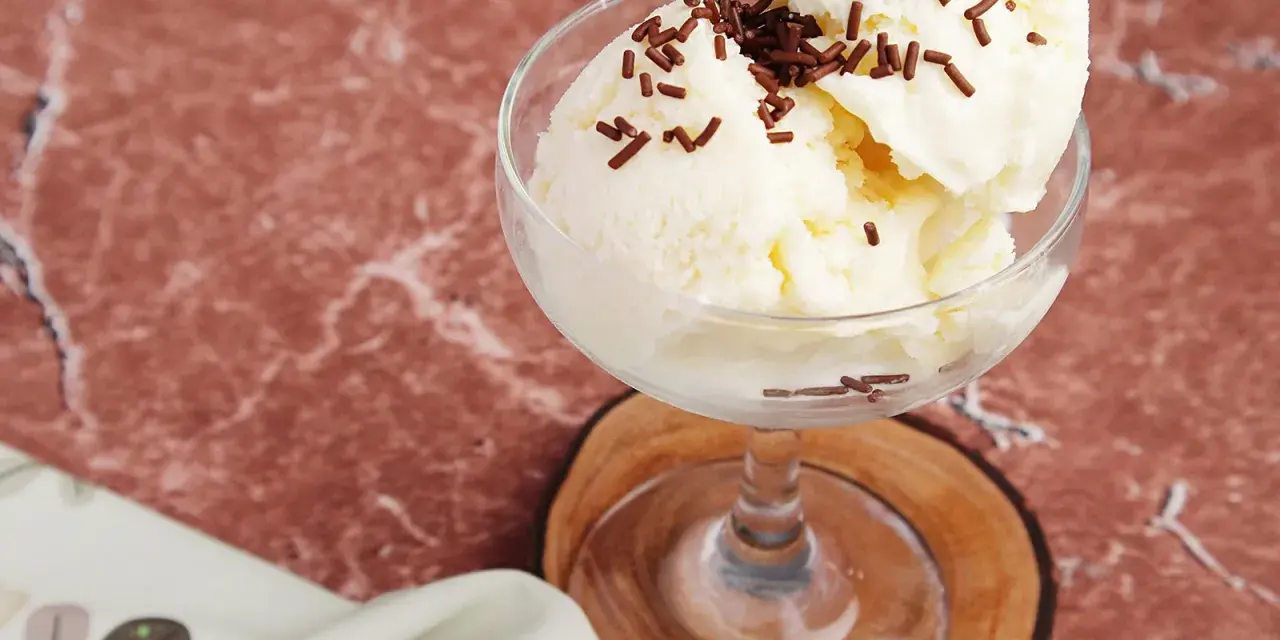 Cremiges Litschi Eis mit Schokostreuseln in einem Glas auf einem Holzuntersetzer.