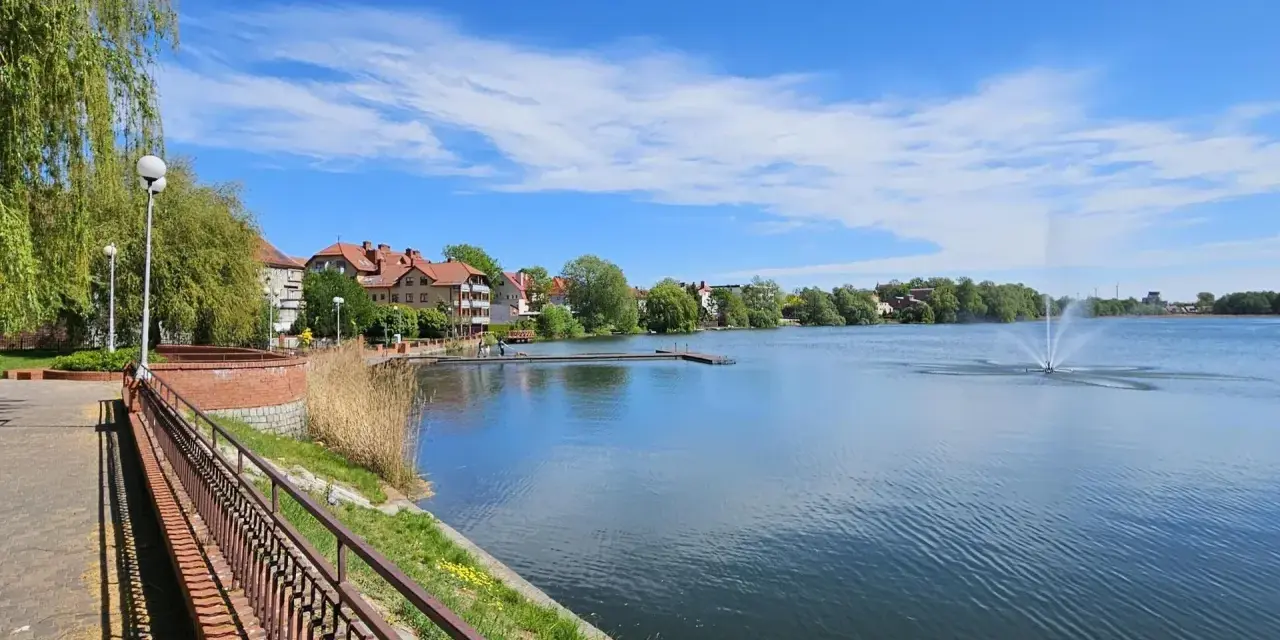 Spokojne jezioro zamkowe z fontanną i drewnianym pomostem, otoczone zielenią i zabudowaniami.