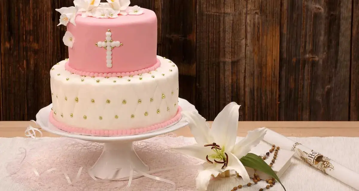 Elegancki dwupoziomowy tort na chrzest w różowo-białych barwach, ozdobiony krzyżem i kwiatami. Obok lilia, różaniec i świeca.