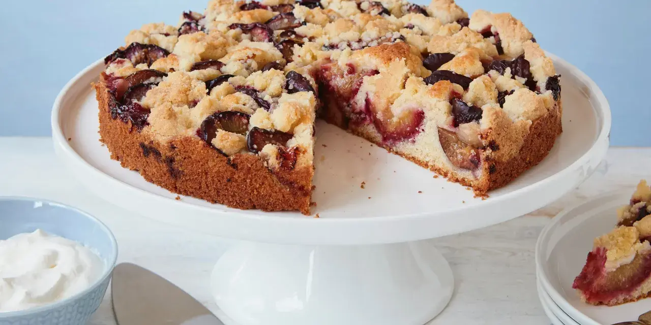 Ein einfacher Pflaumenkuchen mit Streuseln auf einem Tortenständer, daneben eine Schüssel Schlagsahne und ein Tortenheber.
