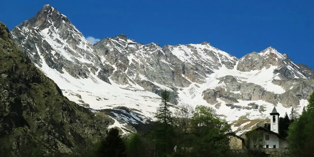 Imponente montagna sacra innevata, con un piccolo campanile bianco che emerge tra gli alberi verdi in primo piano.