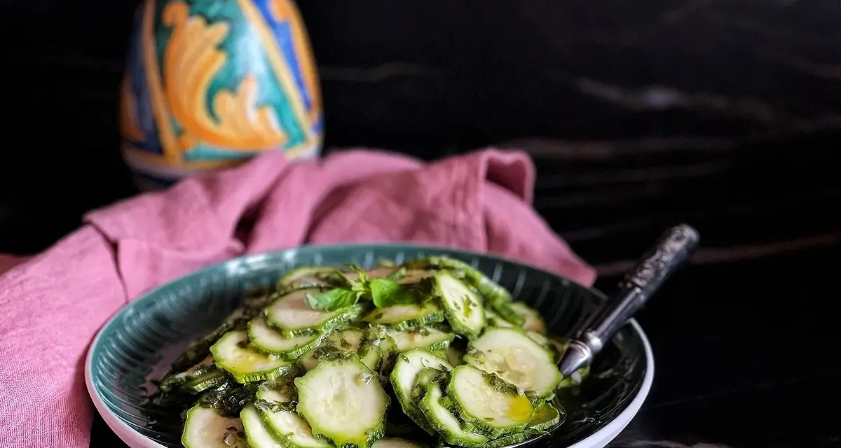 Zucchine marinate cotte, condite con erbe aromatiche, servite su un piatto verde. Sullo sfondo, un oggetto decorativo colorato e un tovagliolo rosa.