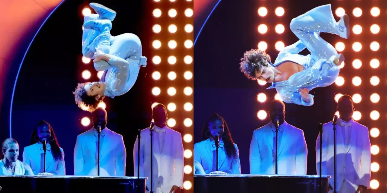 Wokalista w błyszczącym niebieskim stroju wykonuje akrobacje na scenie podczas gali nagród Grammy. Laureaci nagród grammy w białych garniturach stoją za fortepianami.
