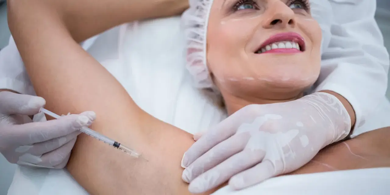 Kobieta z uśmiechem na twarzy poddaje się zabiegowi botox pod pachy. Lekarz w rękawiczkach wykonuje zastrzyk.