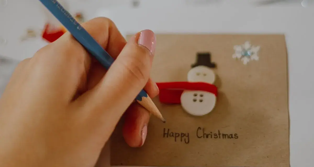 Ręka z ołówkiem pisze "Happy Christmas" na kartce z bałwankiem z guzików. Idealny przykład, jak narysować kartkę świąteczną.