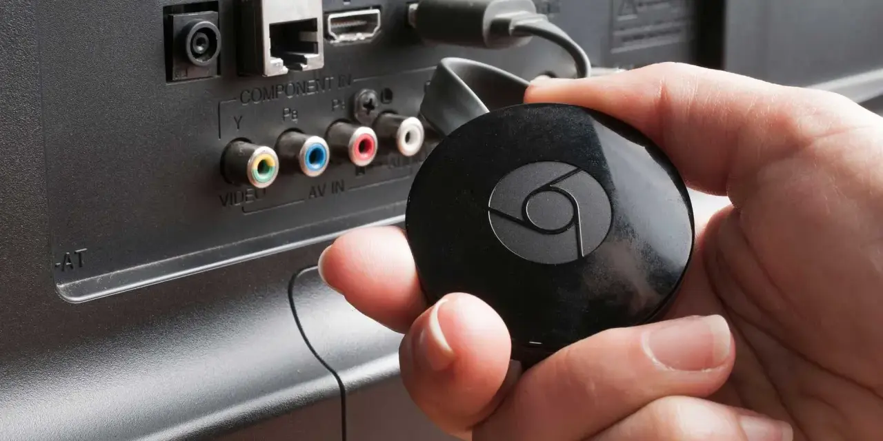 En hand håller en svart Chromecast för att ansluta till en HDMI-port på baksidan av en TV.