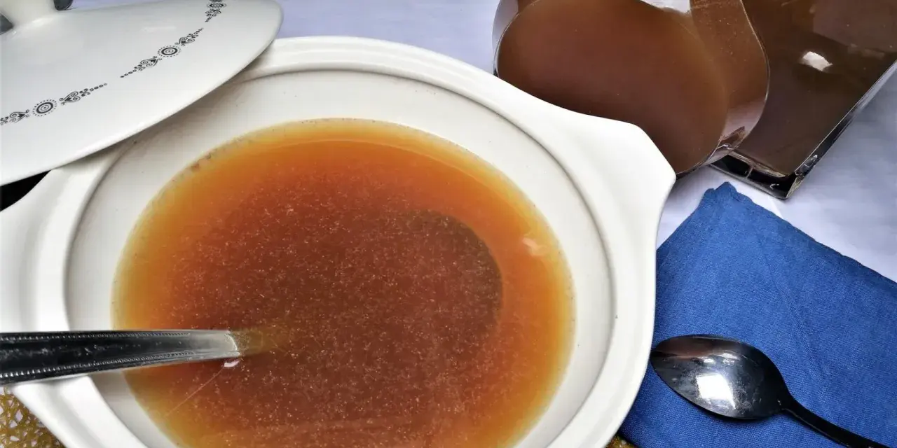 Caldo de carne casero en una cazuela blanca con tapa, junto a una jarra y cucharas.