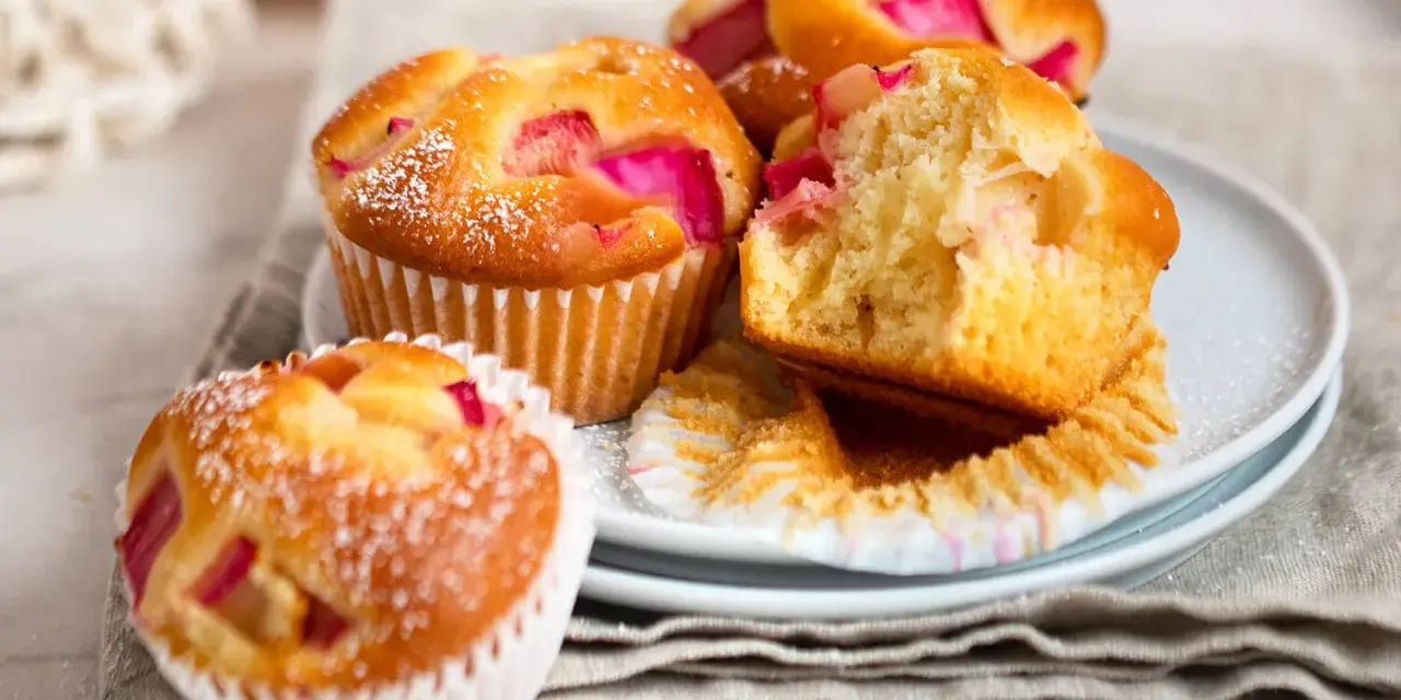 Saftige Rhabarber Muffins, mit Puderzucker bestäubt, auf einem Teller gestapelt. Einer ist angeschnitten und zeigt die rosa Rhabarberstücke.