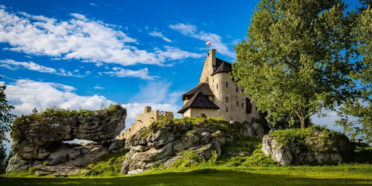 Zamek Bobolice na skale, z polską flagą, pod błękitnym niebem z chmurami. Obok skalny most i zielona trawa.