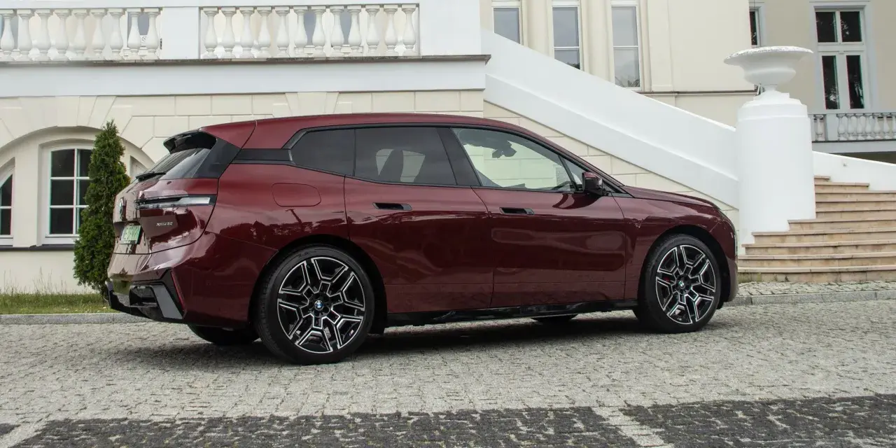 Ciemnoczerwone BMW iX stoi na brukowanym podjeździe przed eleganckim budynkiem.