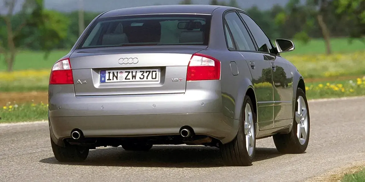 Srebrne Audi B6 sedan z tablicą rejestracyjną IN ZW 370 jedzie po asfaltowej drodze wśród zieleni i żółtych kwiatów.