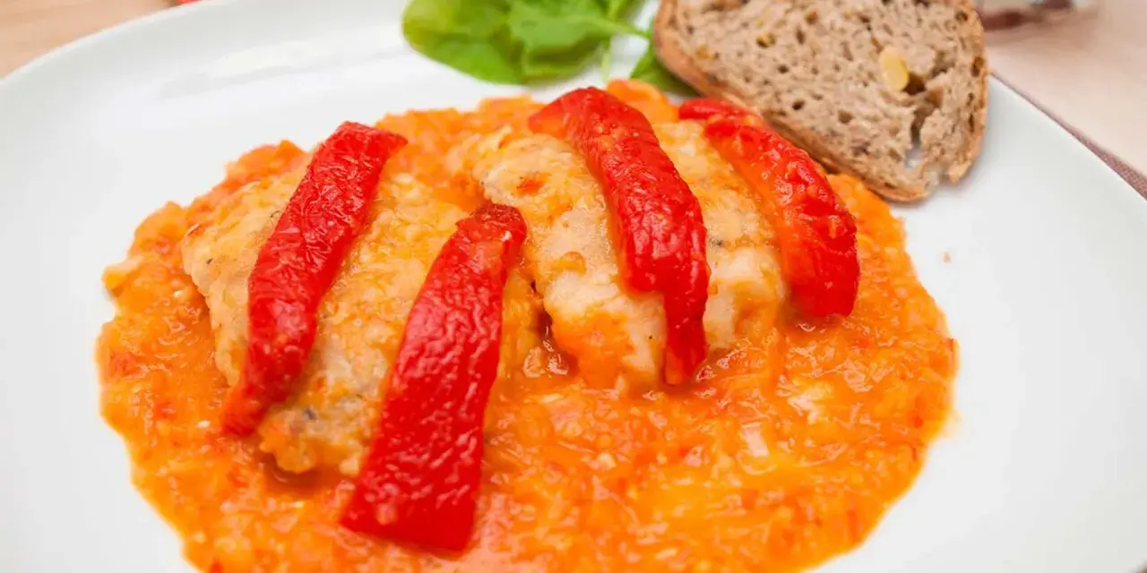 Bacalao a la riojana, un plato tradicional con trozos de pescado blanco cubiertos de pimientos rojos y una salsa de tomate y pimientos. Servido con pan y hojas verdes.