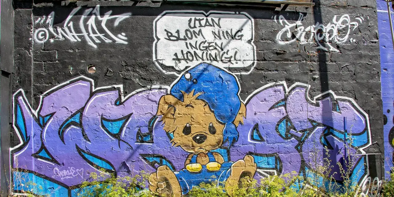 Enkel graffiti på en tegelvägg visar en brun nallebjörn i blå overall och mössa, med texten "Utan blommor ingen honung!".