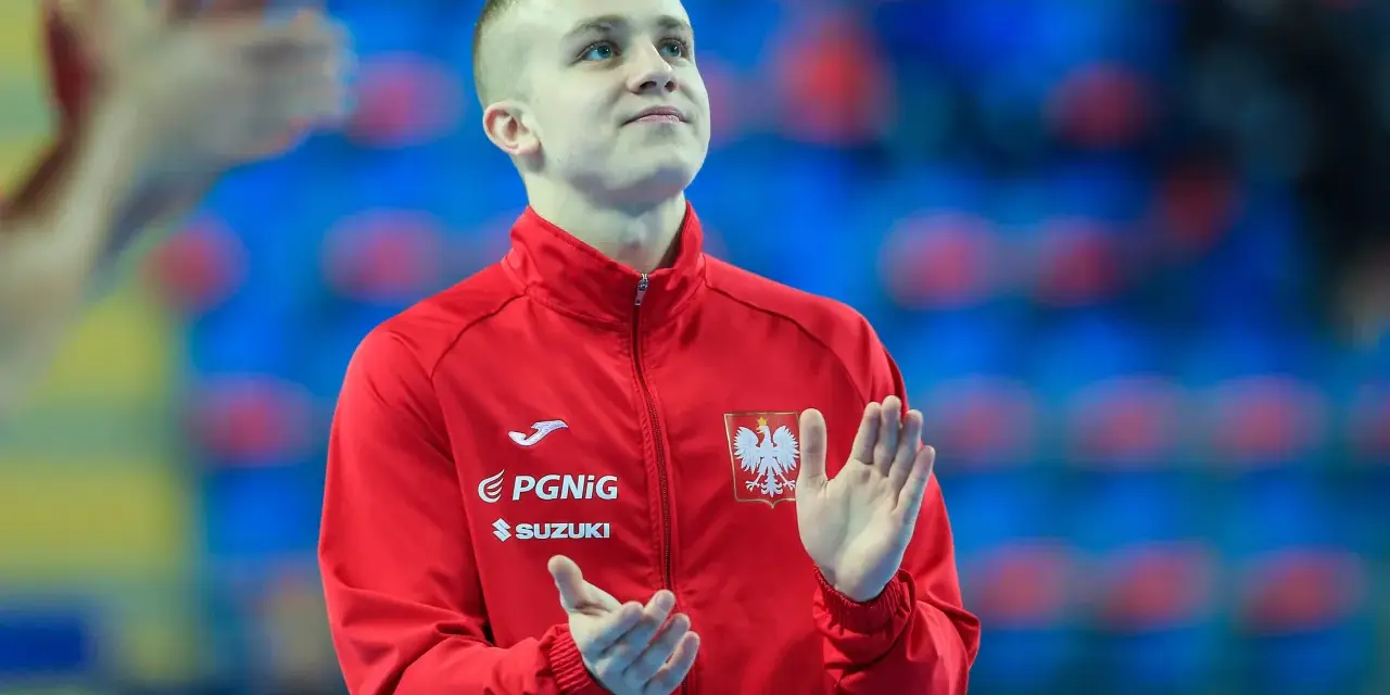 Adam Jędraszek w czerwonej kurtce z orłem, z dłońmi uniesionymi w geście radości lub powitania.