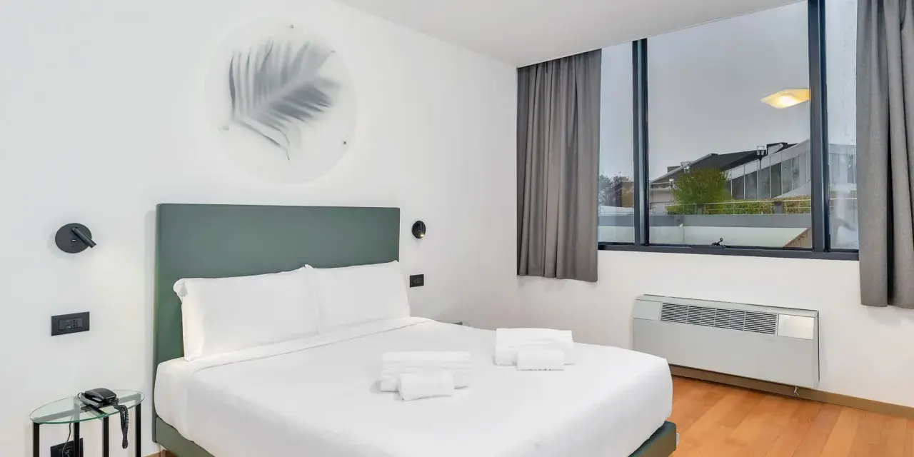 A modern hotel room with a king-size bed, white linens, and a green headboard.