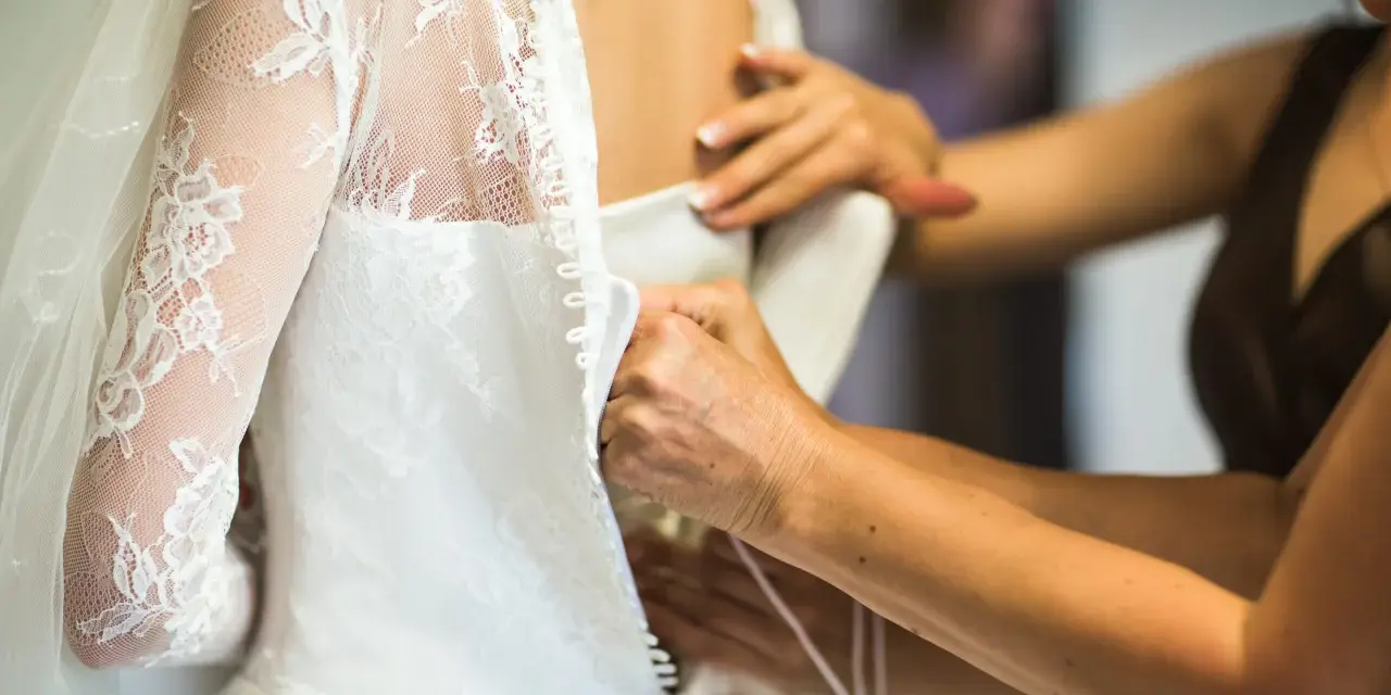 Dettaglio di abiti da sposa riutilizzabili: mani che chiudono bottoncini su un vestito bianco con pizzo e velo.