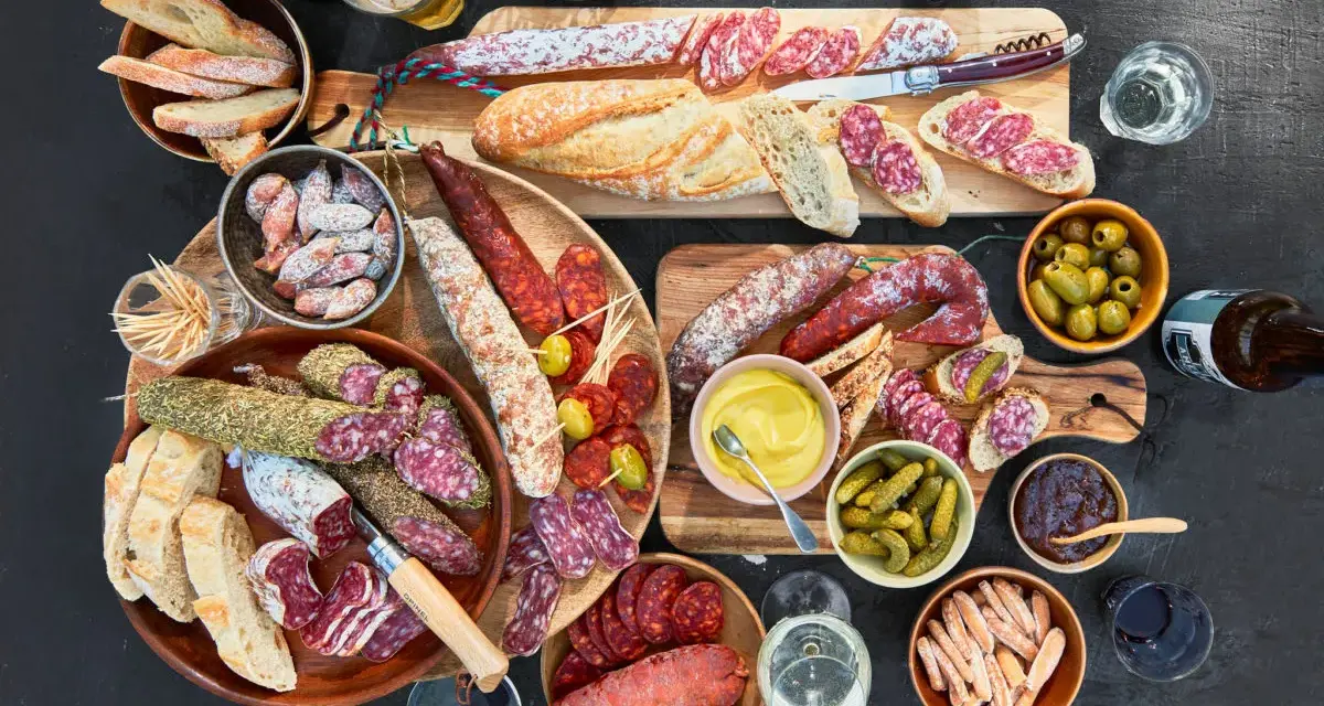 Eine reichhaltige Wurstplatte anrichten mit Baguette, Oliven, Cornichons und Senf.