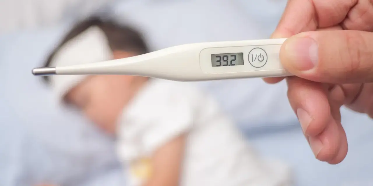 Cyfrowy termometr pokazuje 39.2°C, wskazując wysoką temperaturę ciała u chorego dziecka leżącego w łóżku.