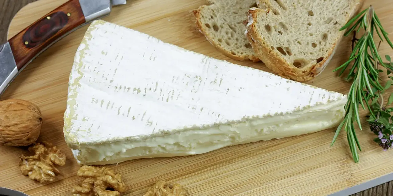 Tranche de fromage à croute lavée crémeux, accompagnée de pain, noix et herbes aromatiques sur une planche en bois.