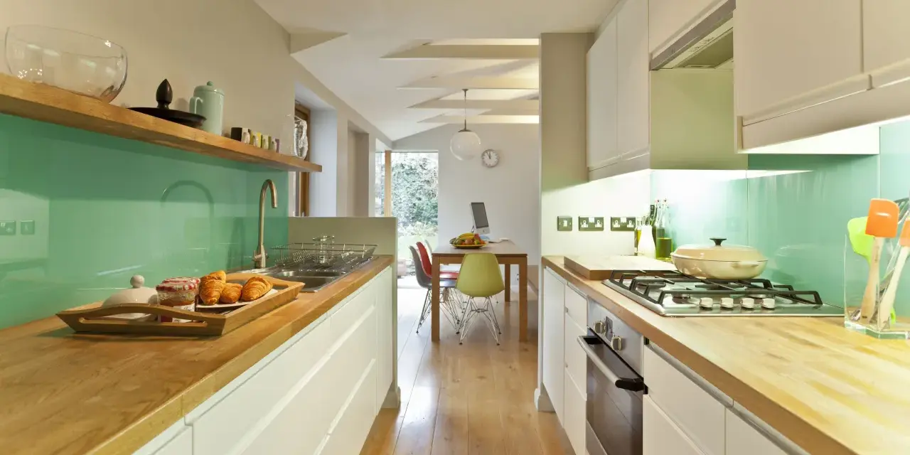 Cocina alargada con encimeras de madera, salpicadero verde turquesa y electrodomésticos integrados. Perfecta para distribuir el espacio.