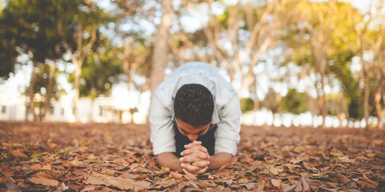 A man in a light blue shirt is in a posture of christian prostration, hands clasped on the ground covered in fallen leaves.