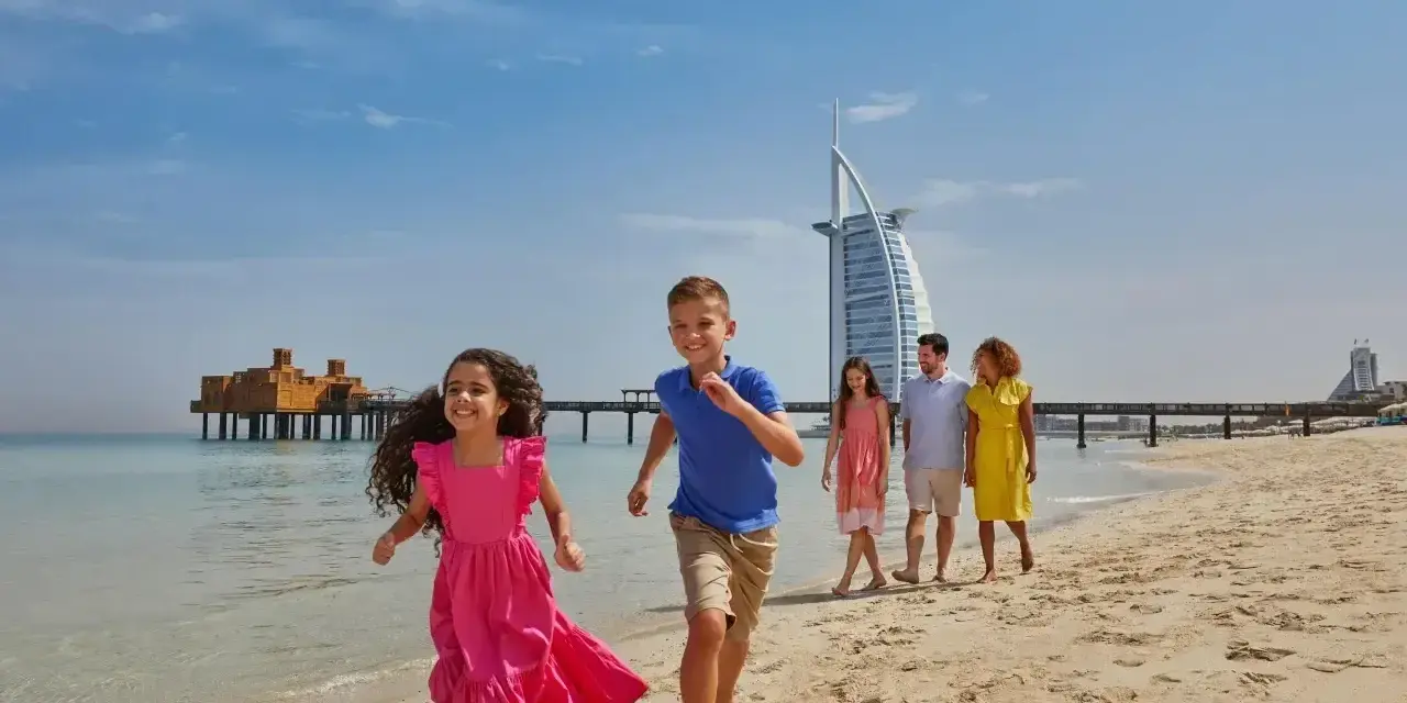 Rodzina cieszy się słonecznym dniem na plaży w Dubaju. Dzieci biegną w stronę morza, a w tle widać słynny hotel Burj Al Arab.