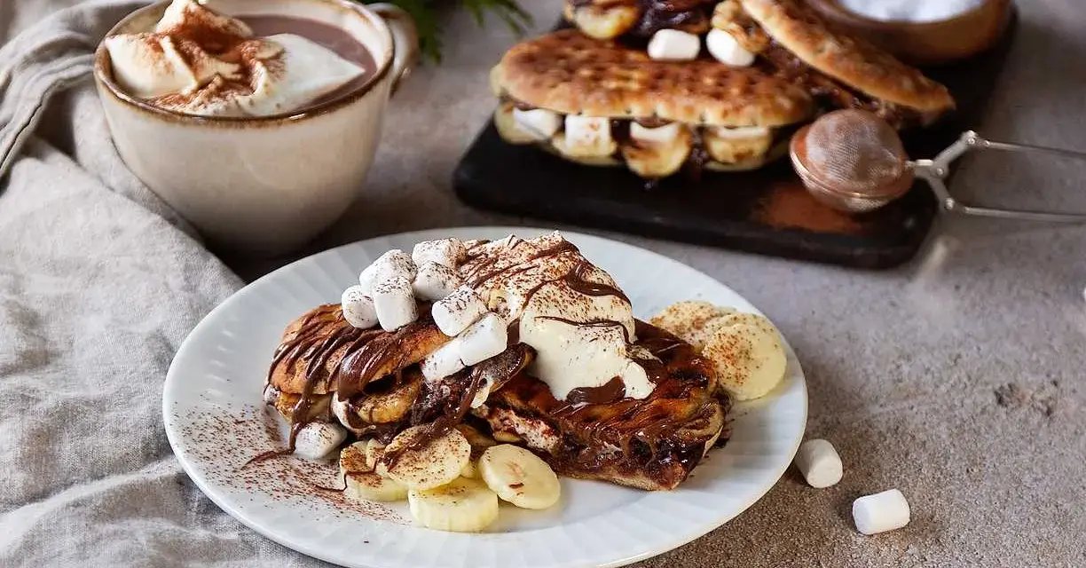 En härlig nutella macka med banan och marshmallows, toppad med grädde och kakao. Bredvid står en kopp varm choklad.