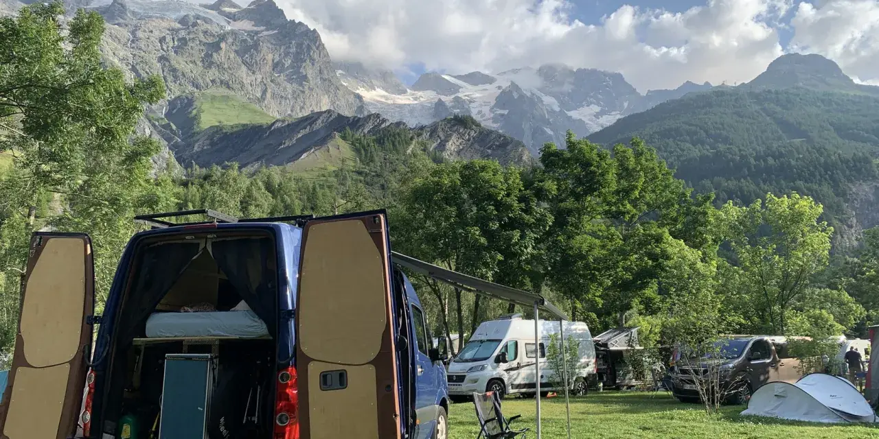 En campingplats med flera husbilar, inklusive en blå med öppna bakdörrar, i en dal med snöklädda berg i bakgrunden. En perfekt plats för äventyr med edins husbilar.
