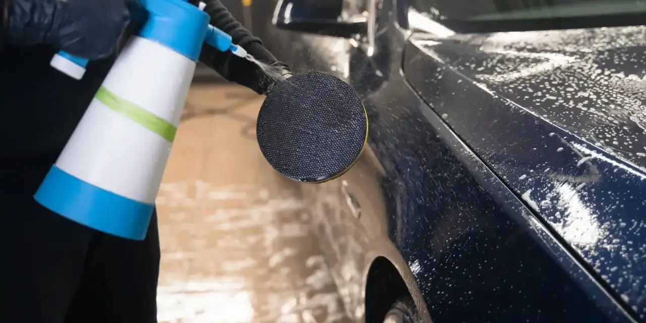 En person med svarta handskar sprutar skum på en mörk metallicbil med en blå och vit sprayflaska. Bilvax metallic ger en glansig finish.