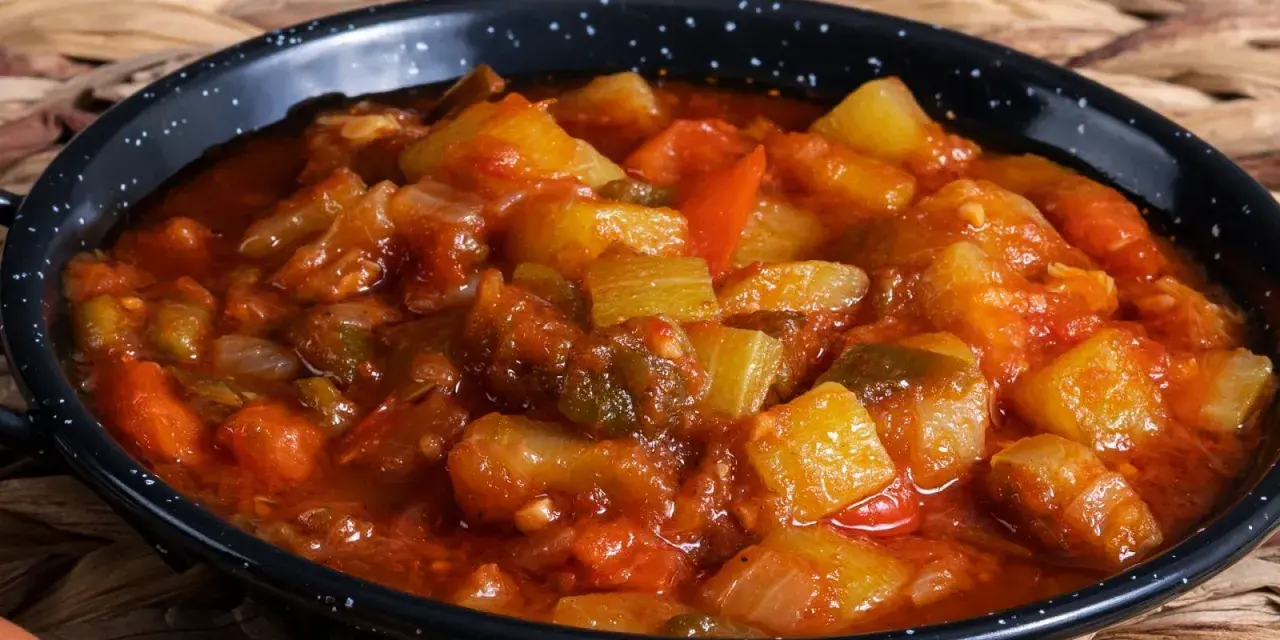 Un plato humeante de pisto manchego, con trozos de calabacín, pimiento y tomate en una salsa rojiza, servido en una cazuela negra.