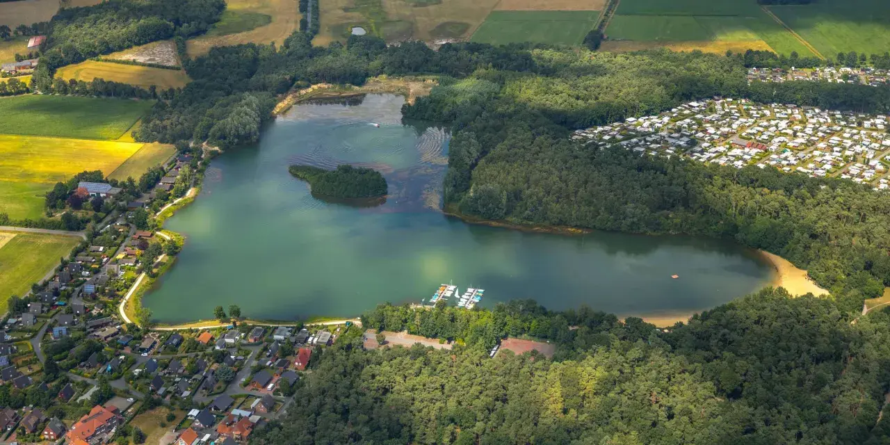 Luftaufnahme des Feldmarksees Sassenberg mit einer kleinen Insel, Booten am Steg, einem Wohngebiet und Feldern.