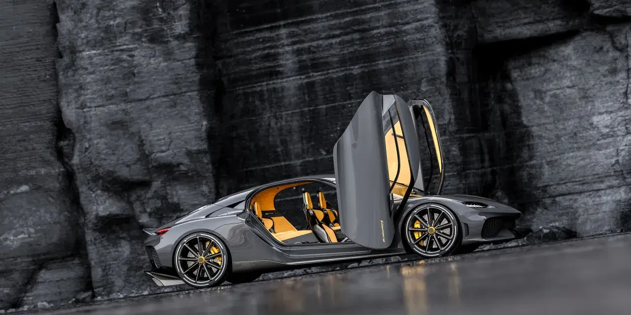 Szary Koenigsegg Gemera, czteromiejscny supersamochód z otwartymi drzwiami, prezentuje swoje żółte wnętrze na tle skalistej ściany.