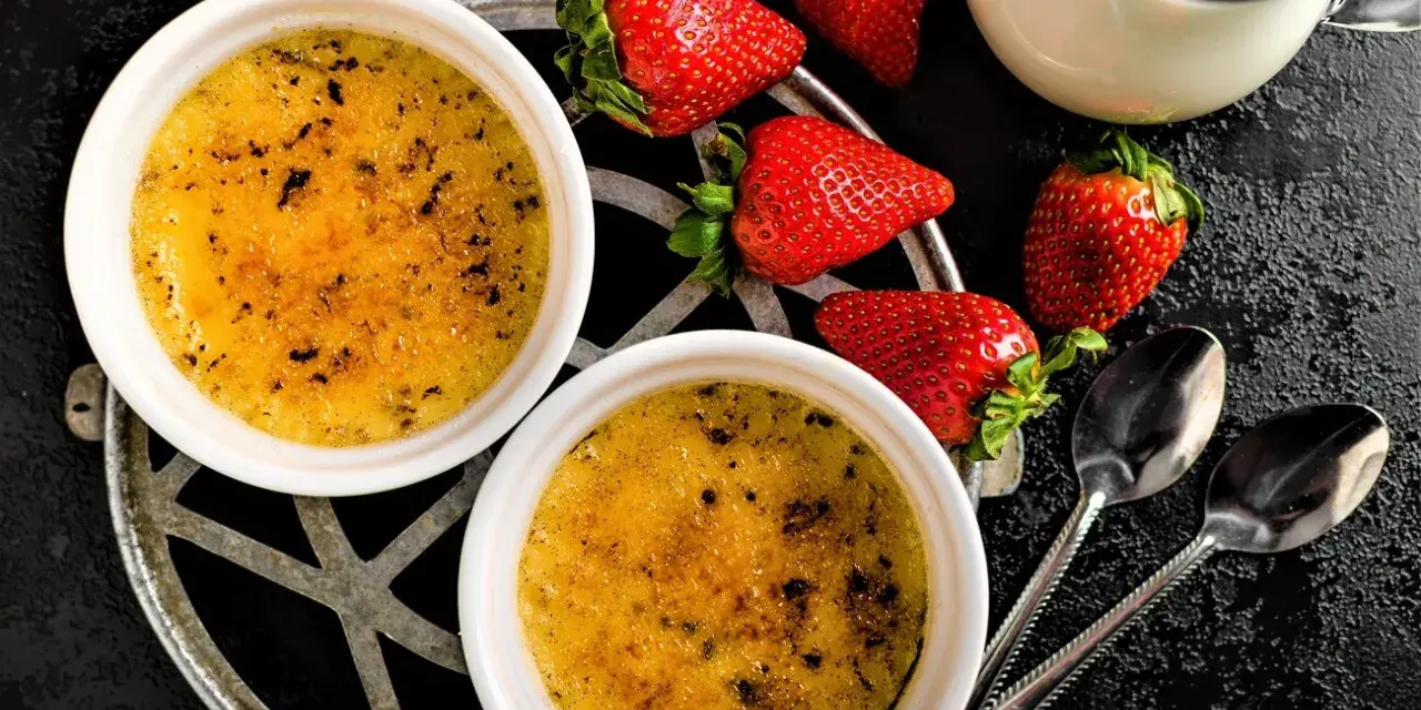 Zwei cremige Crème brûlée mit karamellisierter Kruste, garniert mit frischen Erdbeeren und einem Milchkännchen.