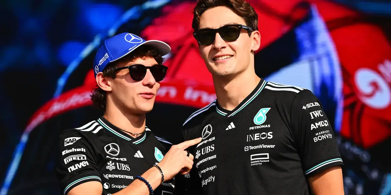 Dos pilotos de Fórmula 1, George Russell y un compañero, sonríen con gafas de sol. Russell lleva una gorra azul y ambos visten camisetas negras con logotipos de patrocinadores.
