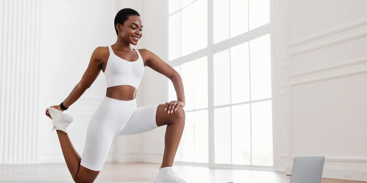 Femme en tenue de sport blanche effectuant un exercice quadriceps sur un tapis, guidée par un ordinateur portable.