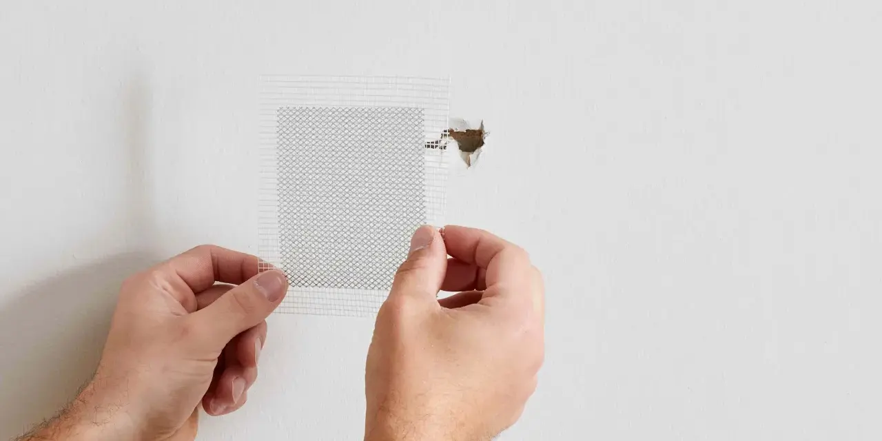 Hands apply a mesh patch to a hole in a white wall, demonstrating how to patch a hole in the wall.