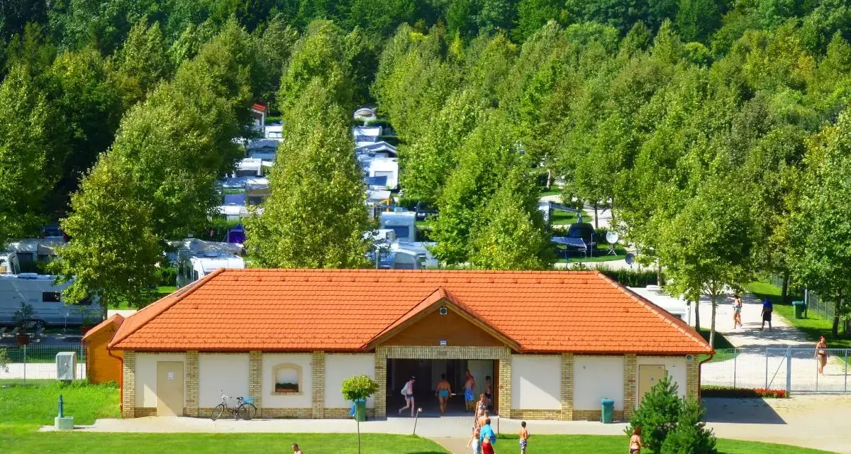 Słoneczny dzień na kempingu Liptot. Ludzie odpoczywają na trawie, a w tle widać rzędy kamperów i przyczep kempingowych wśród drzew.