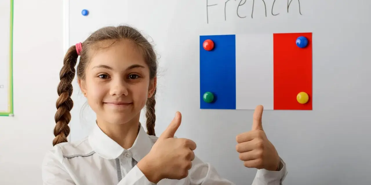 Uśmiechnięta dziewczynka z warkoczami pokazuje kciuki w górę na tle tablicy z napisem "French" i flagą Francji. To symbol pozytywnego podejścia do nauki, być może w ramach systemu edukacji we Francji.