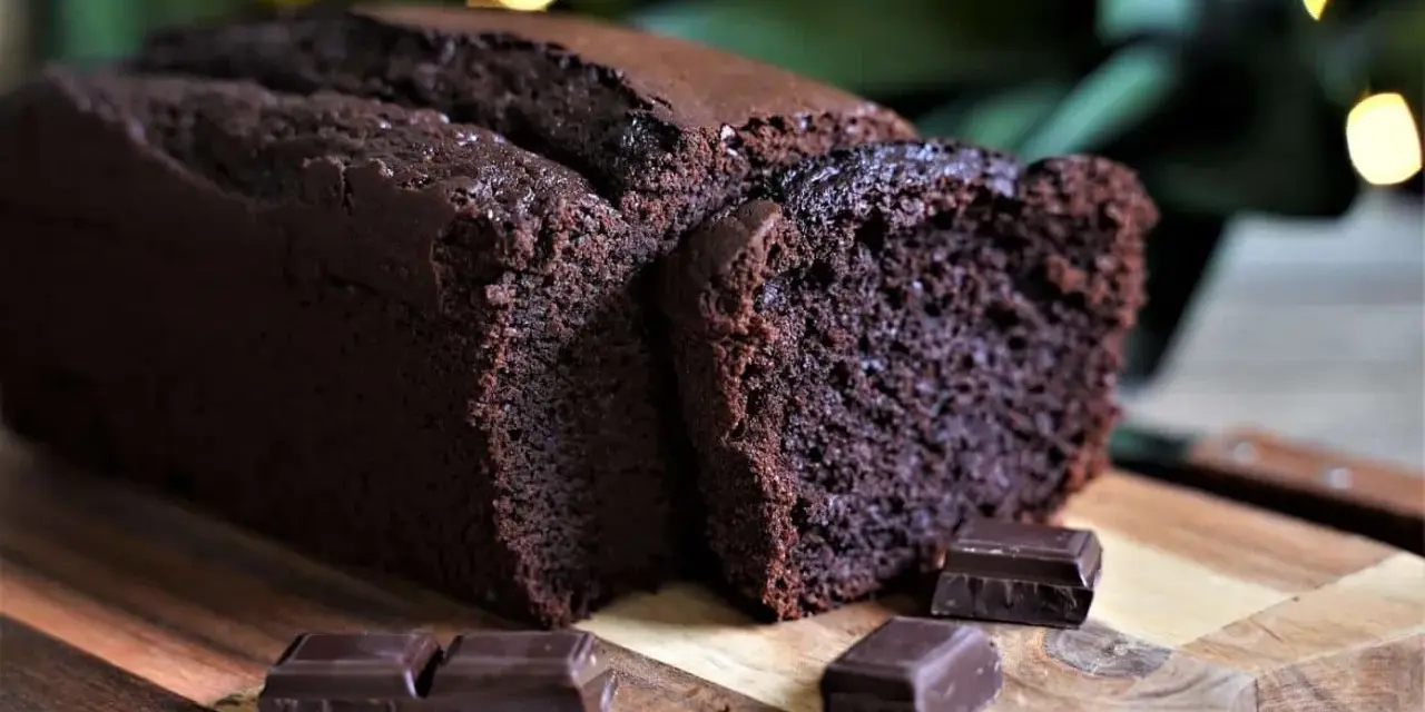 Un délicieux cake au chocolat, moelleux et intense, coupé en tranches sur une planche en bois, avec des carrés de chocolat noir en garniture.