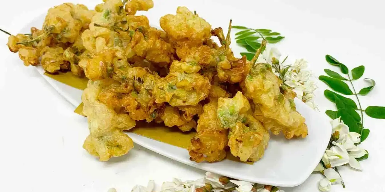 Fiori di acacia fritti, dorati e croccanti, serviti su un piatto bianco con rametti di fiori e foglie fresche.