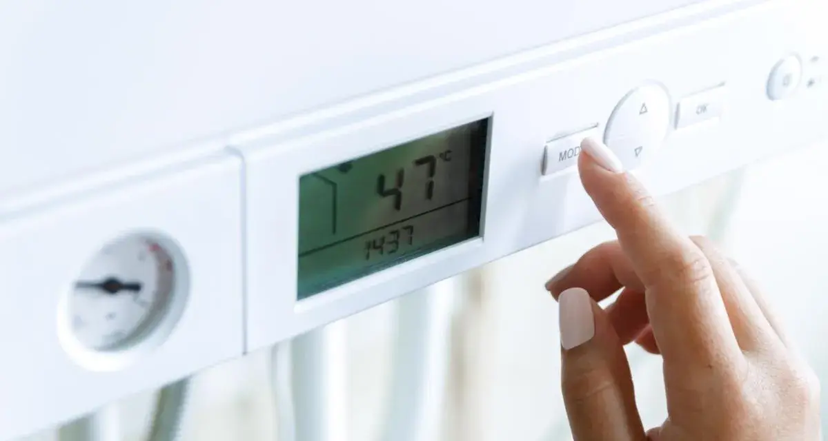 Dito, la mano preme il pulsante "MOD" su una caldaia. Il display mostra 47°C, ma la pressione acqua calda è bassa.