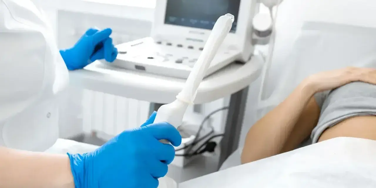 Lekarz w niebieskich rękawiczkach trzyma sondę USG, przygotowując się do badania. Na ekranie widoczne są prawidłowe wymiary macicy w usg.