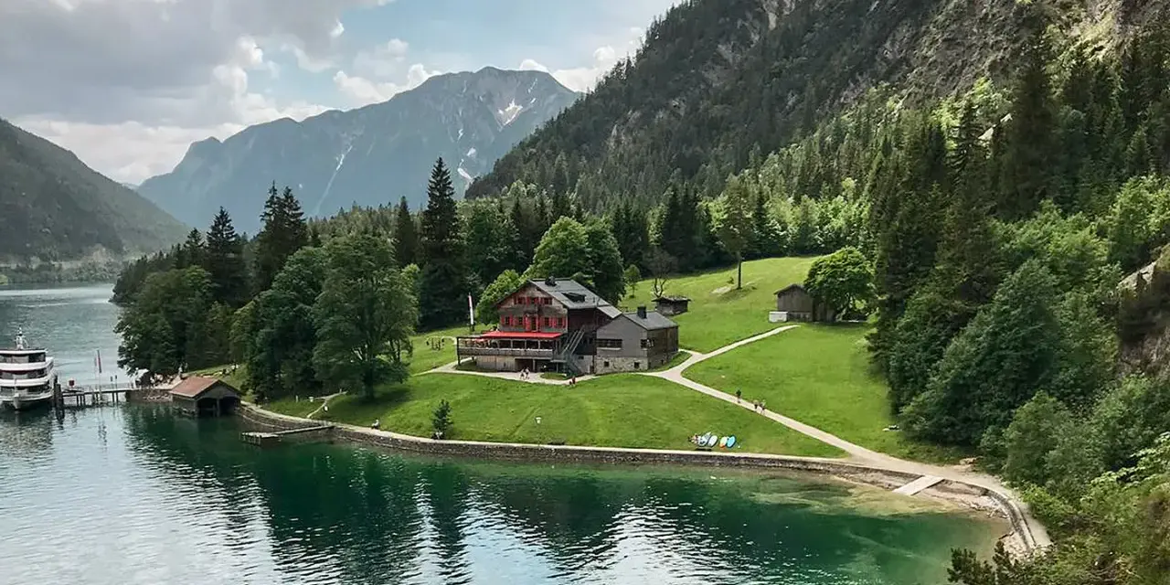 Gaisalm am Achensee: ein Gasthaus mit roten Fensterläden, umgeben von grünen Wiesen und Bergen. Ein Schiff liegt am Steg.