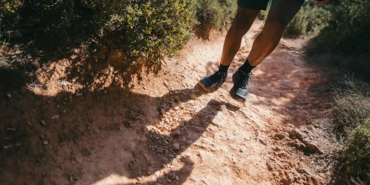 Jambes d'un homme en short et chaussures de trail, s'engageant sur un sentier rocailleux et poussiéreux. L'ombre de son corps s'étire sur le chemin, suggérant un rythme de fast hiking sous le soleil.